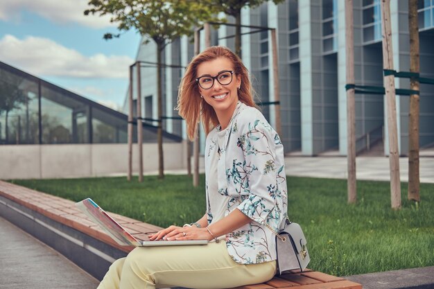 Fröhlicher, stilvoller Modeblogger, der sich im Freien entspannt, am Laptop arbeitet und auf einer Bank gegen einen Wolkenkratzer sitzt.