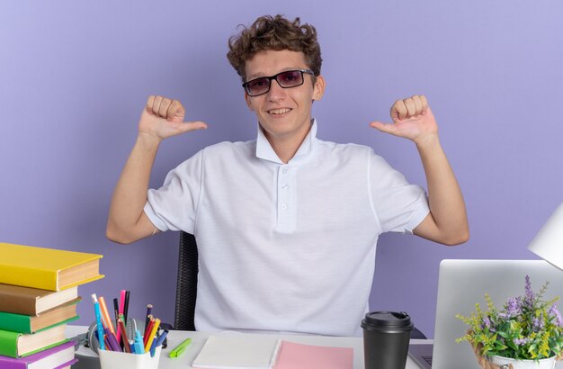 Fröhlicher Student in weißem Poloshirt mit Brille am Tisch sitzend mit Büchern, die selbstbewusst lächeln und auf sich selbst auf blauem Hintergrund zeigen