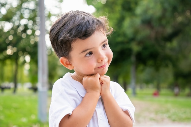 Fröhlicher süßer kleiner Junge, der im Sommerpark steht und posiert, Kinn auf Hände stützt, lächelt und wegschaut. Nahaufnahme. Kindheitskonzept