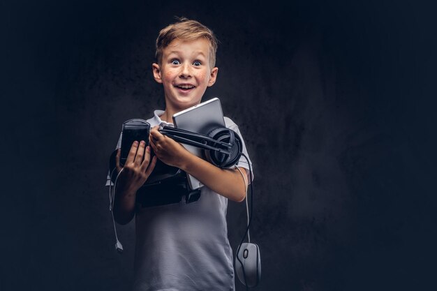 Fröhlicher süßer Schuljunge in einem weißen T-Shirt, hält ein komplettes digitales Set zur Unterhaltung in einem Studio. Getrennt auf einem dunklen strukturierten Hintergrund.