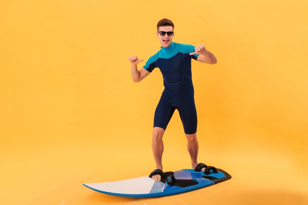 Fröhlicher Surfer in Neoprenanzug und Sonnenbrille mit Surfbrett zeigt sich und schaut in die Kamera