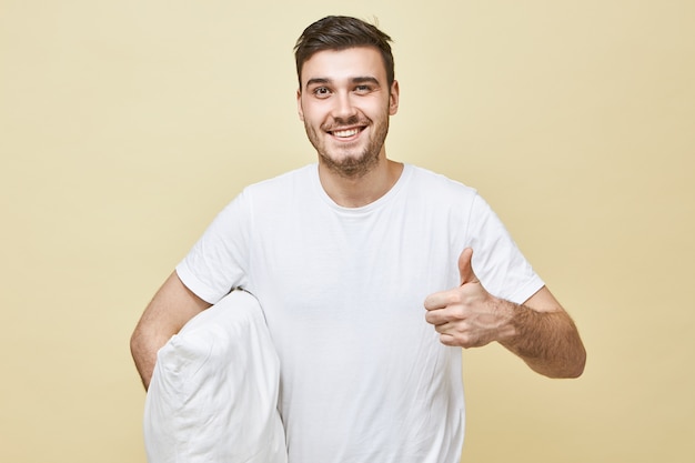 fröhlicher überglücklicher junger kaukasischer Mann im weißen T-Shirt mit strahlendem Lächeln, der sich nach gutem Tiefschlaf auf neuem Kissen entspannt und enegetisch fühlt und Daumen hoch zeigt