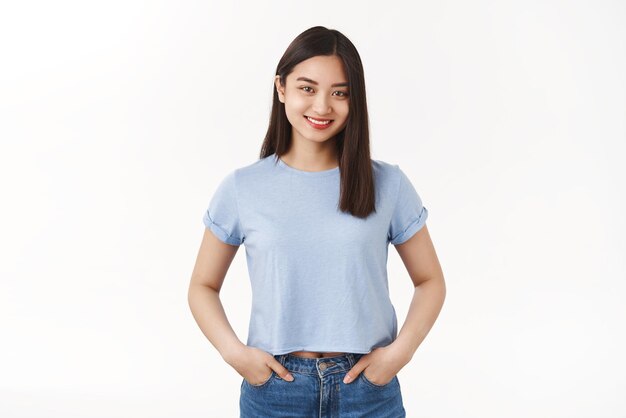 Fröhliches, angenehmes, freundliches, durchsetzungsfähiges, abgehendes asiatisches junges Mädchen, das Hände hält, Taschen, Jeans, die breit lächeln, möchte helfen, hellblaues T-Shirt zu tragen, entspannte, sorglose Pose, die auf weißem Hintergrund steht