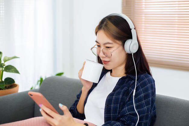 Fröhliches asiatisches Brillenmädchen, das Musik vom Kopfhörer hört und Kaffee trinkt Wohnzimmer Konzept für die Erholung zu Hause