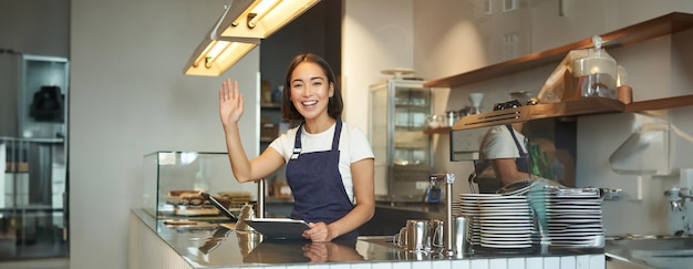 Fröhliches asiatisches Mädchen Barista Barkeeper hinter der Theke hebt den Arm und winkt den Kunden bei der Verarbeitung