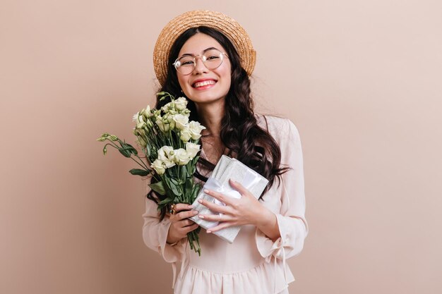 Fröhliches asiatisches Mädchen mit Geschenk und Blumen Lächelnde chinesische Frau mit Hut, die mit Eustoma-Bouquet posiert