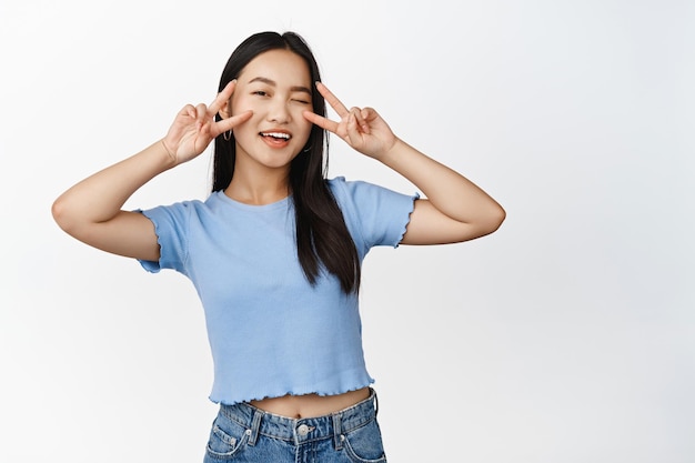 Fröhliches asiatisches Mädchen zwinkert und zeigt vsigns kawaii Geste in der Nähe von Augen, die Sie grüßen, wenn Sie im T-Shirt auf weißem Hintergrund stehen
