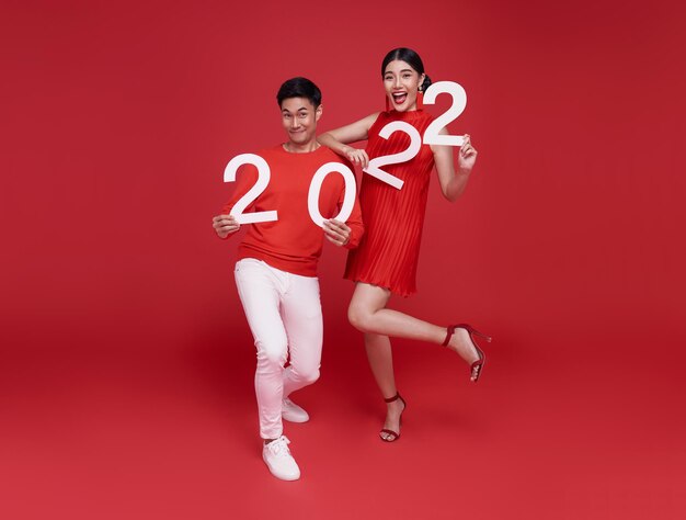 Fröhliches asiatisches Paar in roter Freizeitkleidung mit der Nummer 2022, das ein frohes neues Jahr mit einem Lächeln auf hellrotem Hintergrund begrüßt.