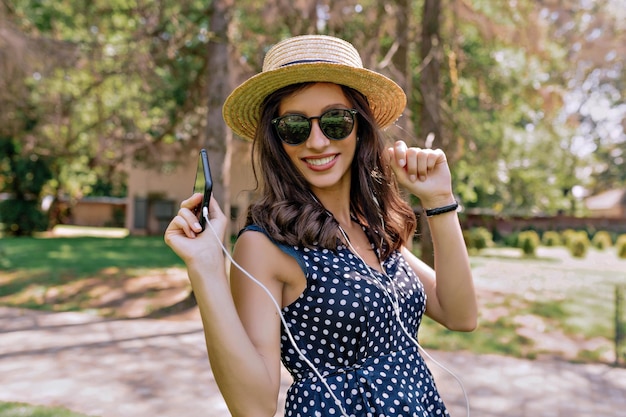 Fröhliches, aufgeregtes, hübsches Mädchen mit dunklem Haar und gebräunter Haut, das eine Sonnenbrille und einen Sommerhut trägt, hört Musik im Kopfhörer und tanzt