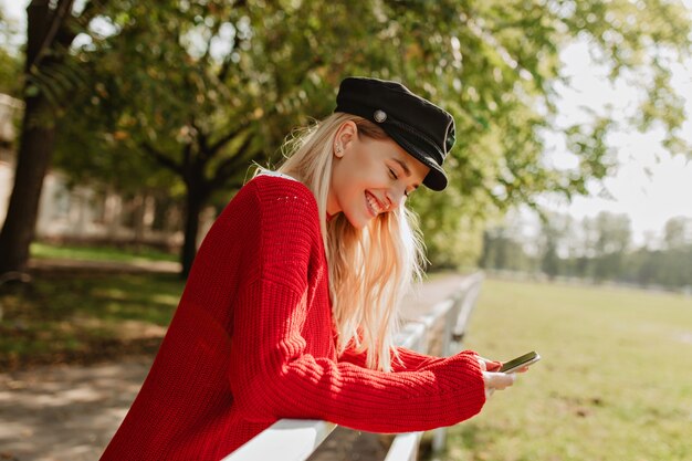 Fröhliches blondes Mädchen, das glücklich im Herbstpark lächelt. Hübsche Frau, die ihr Telefon unter gelben Bäumen betrachtet.