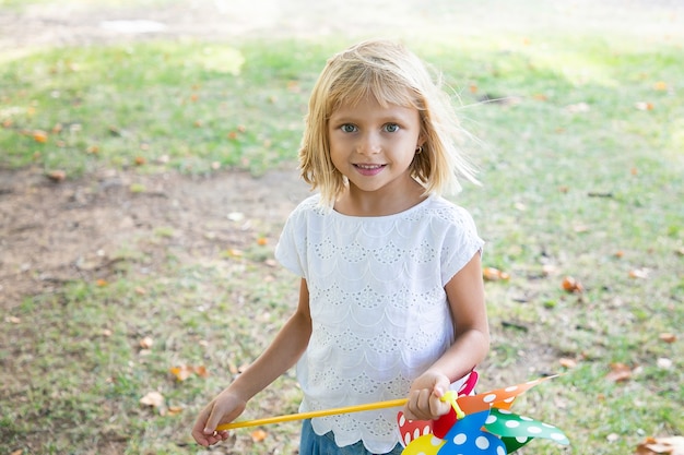 Fröhliches blondes Mädchen, das im Park spielt, Windrad hält und lächelt. Vorderansicht. Outdoor-Aktivitätskonzept für Kinder