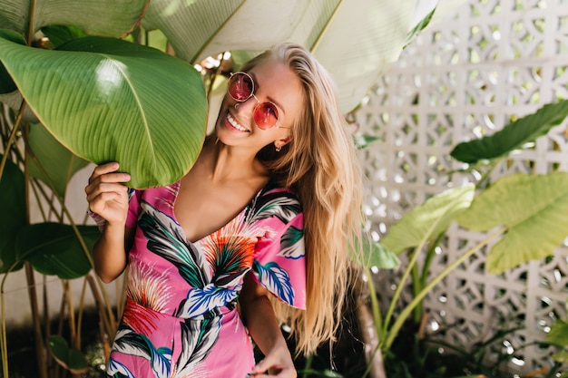 Fröhliches blondes Mädchen, das mit aufrichtigem Lächeln nahe großem grünem Blatt aufwirft.