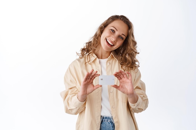 Fröhliches blondes Mädchen kippt den Kopf und lächelt, zeigt Plastikkreditkarte über der Brust, empfiehlt ein Bankangebot, weißer Hintergrund