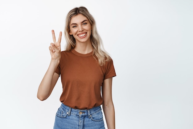 Fröhliches blondes Mädchen zeigt Frieden, V-Zeichen-Geste und Lächeln und steht in der Nähe von leerem Kopierraum für Werbung auf weißem Studio