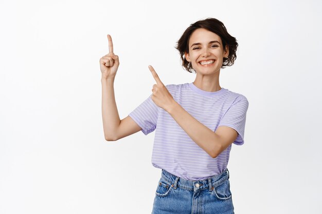 Fröhliches brünettes Mädchen, das mit den Fingern auf die obere linke Ecke zeigt und stolz im T-Shirt auf Weiß steht.