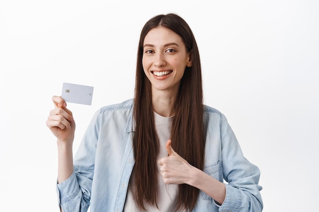 Fröhliches brünettes Mädchen zeigt Kreditkarte und Daumen hoch, empfiehlt Bank, lobt kontaktlose einfache Zahlung, gute Rabatte, steht auf weißem Hintergrund