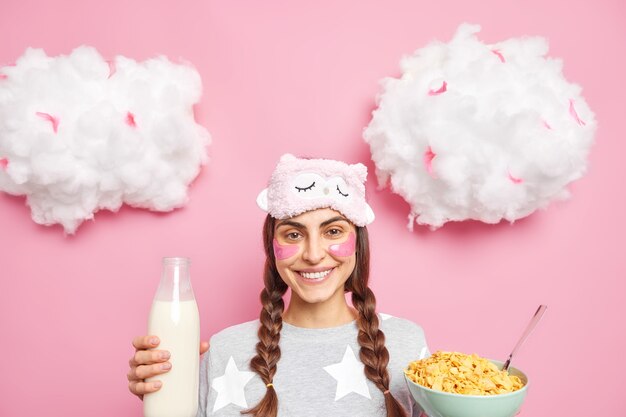 Fröhliches dunkelhaariges Mädchen mit gekämmten Zöpfen hält eine Schüssel mit Müsli und Milch trägt Pyjama