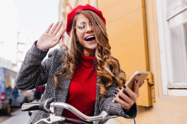 Fröhliches europäisches weibliches Modell in der lustigen roten Baskenmütze, die glückliche Gefühle ausdrückt und um Straße reitet