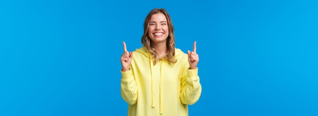 Kostenloses Foto fröhliches, glücklich lächelndes mädchen mit blonden haaren in gelbem hoodie, das mit den fingern nach oben zeigt und lacht