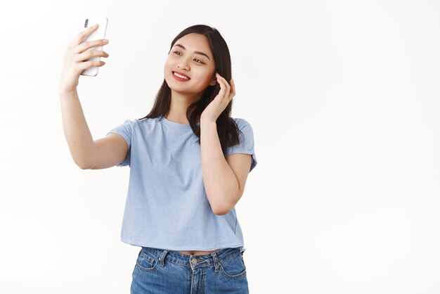Fröhliches, glückliches, selbstbewusstes asiatisches Mädchen, das sich selbst akzeptiert, eigene Erscheinung überprüfen, Haarschnitt schauen, Smartphone-Kamera lächelnd, erfreut, Selfie halten, Telefon-Videoanruf, Freund, weißer Hintergrund