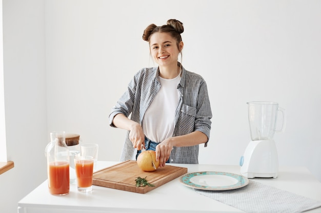 Fröhliches hübsches Mädchen lächelnd, das Grapefruit zum Frühstück über weißer Wand schneidet. Gesunder Detox-Smoothie. Fitness Essen.