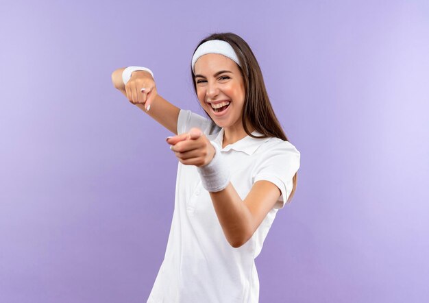 Fröhliches hübsches sportliches Mädchen mit Stirnband und Armband, das Sie isoliert auf lila Wand gestikuliert