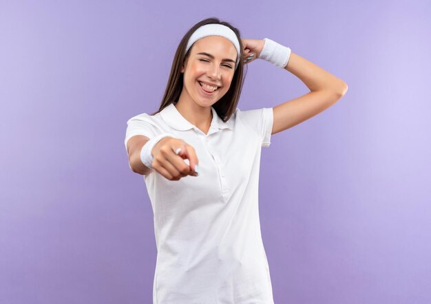 Fröhliches hübsches sportliches Mädchen mit Stirnband und Armband, das starkes Zwinkern zeigt und Zunge isoliert auf lila Wand zeigt