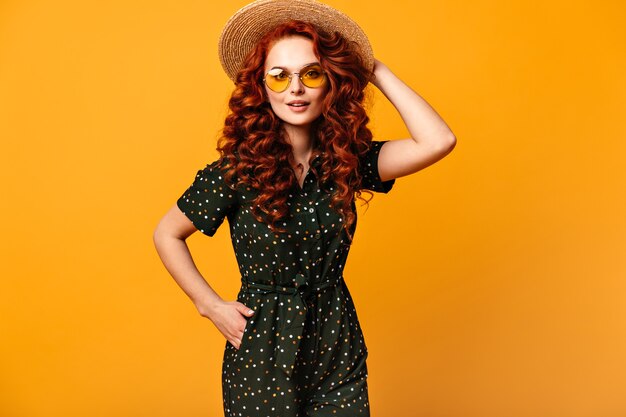 Fröhliches Ingwermädchen in der Sonnenbrille, die mit Hand in der Tasche aufwirft. Studioaufnahme der raffinierten kaukasischen Dame im Strohhut lokalisiert auf gelbem Hintergrund.