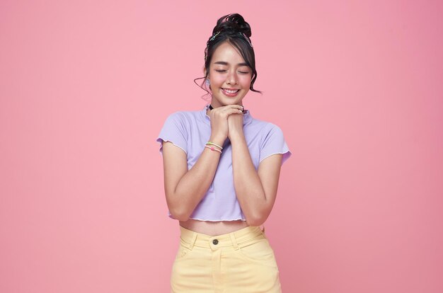 Fröhliches junges asiatisches Teenager-Mädchen, das Händchen hält, betet in der Nähe des Mundes auf rosa Hintergrund