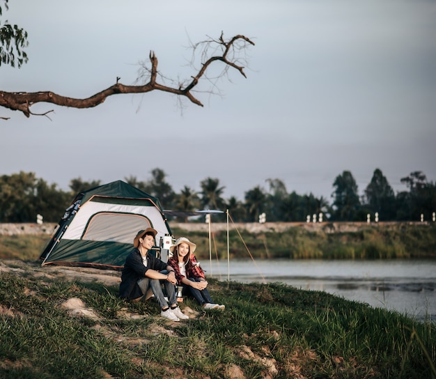 Fröhliches junges Backpacker-Paar, das vor dem Zelt in der Nähe des Sees mit Kaffeesatz sitzt und während des Campingausflugs im Sommerurlaub frische Kaffeemühle macht