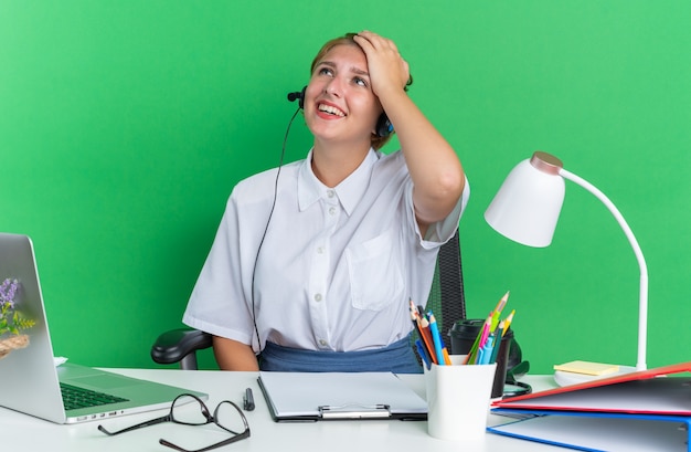 Kostenloses Foto fröhliches junges blondes callcenter-mädchen mit headset, das am schreibtisch mit arbeitswerkzeugen sitzt und die hand auf dem kopf hält, isoliert auf grüner wand nach oben schauend