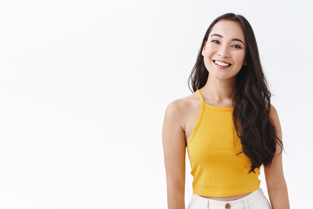 Fröhliches junges brünettes asiatisches Mädchen in stylischem gelbem Top genießen Urlaub im Ausland sonnigen Urlaub lächelnd und lachend glücklich stehend entspannt in lässiger Chill-Pose weißen Hintergrund