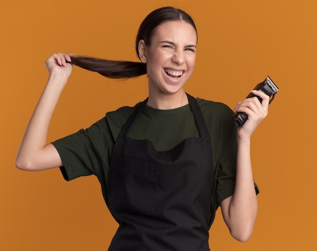 Fröhliches junges brünettes Friseurmädchen in Uniform hält ihre Zopf- und Haarschneidemaschine isoliert auf oranger Wand mit Kopierraum