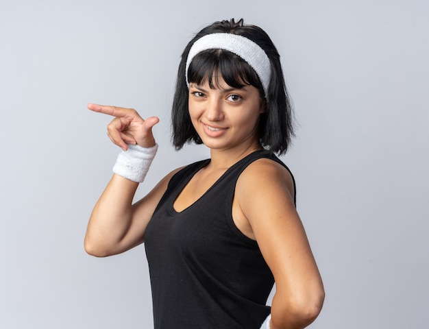 Fröhliches junges Fitness-Mädchen mit Stirnband, das die Kamera ansieht und selbstbewusst lächelt und mit dem Zeigefinger auf die Seite zeigt, die über Weiß steht