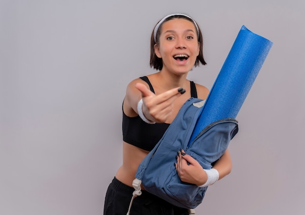 Fröhliches junges Fitnessmädchen in Sportbekleidung hlding Rucksack mit Yogamatte glücklich und positiv machen kommen in Geste über weiße Wand stehen