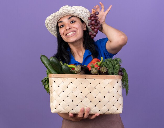 Fröhliches junges kaukasisches Gärtnermädchen in Uniform und Hut, das einen Korb mit Gemüse und Trauben isoliert auf lila Wand hält