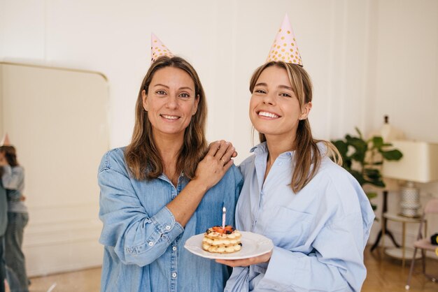 Fröhliches junges kaukasisches Geburtstagskind mit Mutter, die in die Kamera schaut und Kuchen mit Kerze im Zimmer hält. Urlaubskonzept