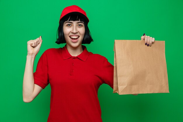 Fröhliches junges kaukasisches Liefermädchen, das Lebensmittelverpackungen hält und ihre Faust hochhält