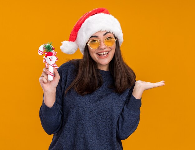 Fröhliches junges kaukasisches Mädchen in Sonnenbrille mit Weihnachtsmütze hält Zuckerstange und hält die Hand isoliert auf oranger Wand mit Kopierraum offen open