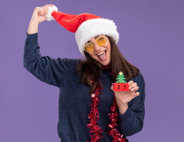 Fröhliches junges kaukasisches Mädchen in Sonnenbrille mit Weihnachtsmütze und Girlande um den Hals streckt die Zunge heraus und hält Weihnachtsbaumschmuck isoliert auf lila Wand mit Kopierraum