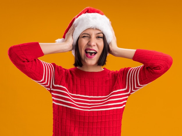 Fröhliches junges Mädchen mit Weihnachtsmütze, das die Kamera anschaut und die Hände auf dem Kopf hält, isoliert auf orangefarbenem Hintergrund