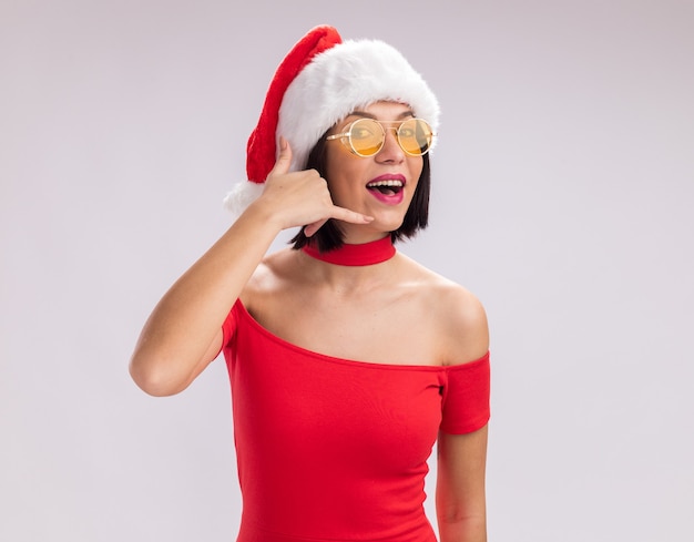 Fröhliches junges Mädchen mit Weihnachtsmütze und Brille, das in die Kamera schaut und die Anrufgeste isoliert auf weißem Hintergrund mit Kopierraum macht