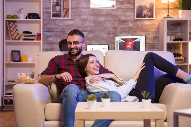 Fröhliches junges Paar lacht, während es nachts eine Fernsehsendung im Fernsehen sieht. Paar sitzt auf dem Sofa.