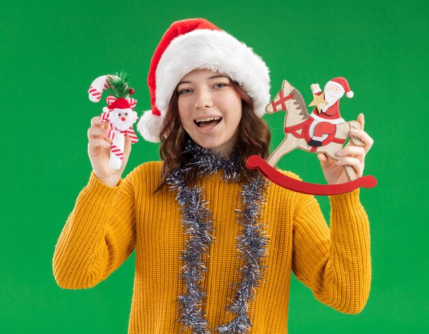 Fröhliches junges slawisches Mädchen mit Weihnachtsmütze und mit Girlande um den Hals, die Zuckerstange und Weihnachtsmann auf Schaukelpferddekoration isoliert auf grüner Wand mit Kopierraum hält