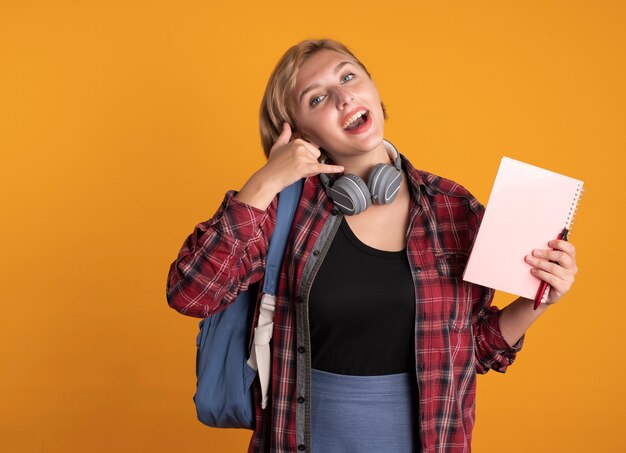 Fröhliches junges slawisches Studentenmädchen mit Kopfhörern, das Rucksack trägt, hält Notizbuch und Stiftgesten rufen mich an