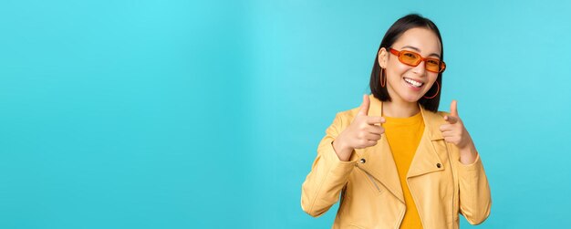 Fröhliches junges stilvolles chinesisches Mädchen mit Sonnenbrille zeigt mit zufriedenem Lächeln mit dem Finger auf die Kamera und wählt Sie aus, um über blauem Hintergrund zu gratulieren