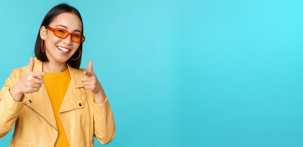 Fröhliches junges stilvolles chinesisches Mädchen mit Sonnenbrille zeigt mit zufriedenem Lächeln mit dem Finger auf die Kamera und wählt Sie aus, um über blauem Hintergrund zu gratulieren