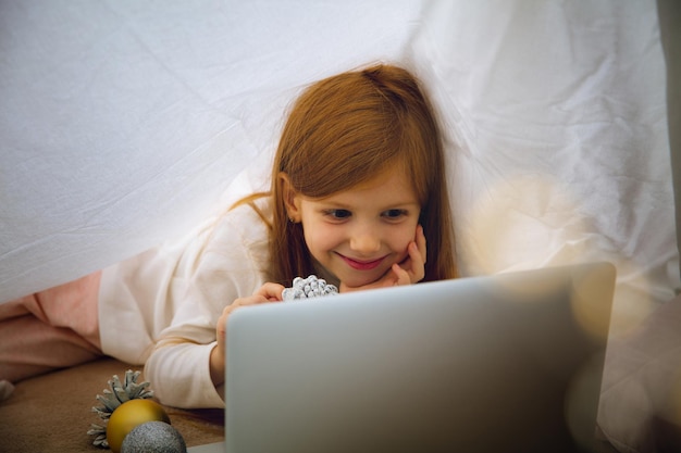 Fröhliches kaukasisches kleines mädchen während eines videoanrufs oder einer nachrichtenübermittlung mit dem weihnachtsmann mit laptop und heimgeräten