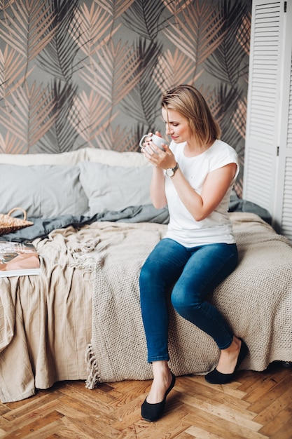 Fröhliches kaukasisches Mädchen mit blondem Haar, weißem T-Shirt, Jeans sitzt im großen hellen Raum und trinkt Kaffee