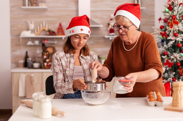 Fröhliches Kind, das traditionelles Teigputting-Mehl in Schüssel mit Sieb macht, das hausgemachtes Lebkuchen-Dessert mit Großmutter kocht, die die Weihnachtszeit feiert. Kind genießt Winterurlaub